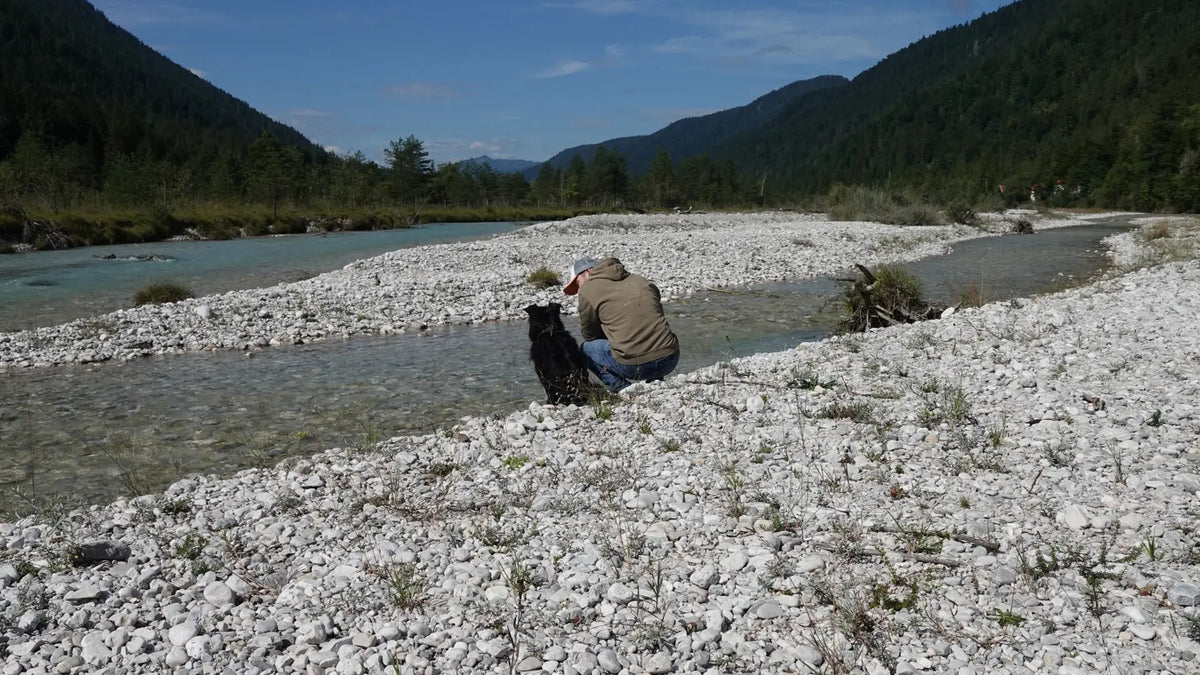 Mann und Hund sitzen gemeinsam an einem klaren Gebirgsfluss inmitten einer bewaldeten Berglandschaft.