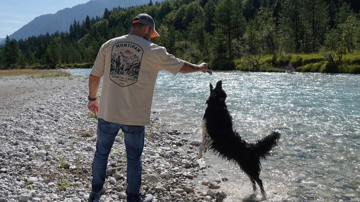 Mann mit Hund an einem Gebirgsfluss trägt das beige Montipaw „The Summit“ Oversized Shirt mit großem Rückenprint.